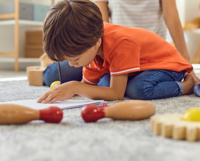 Preschooler Drawing a Picture with Pencils Sitting on Warm Rug with His Brother and Nanny